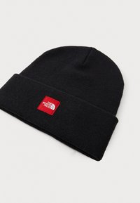 Berretto a maglia nero con risvolto, caratterizzato da una toppa quadrata rossa con lettere bianche e un logo. Texture morbida e design senza cuciture.