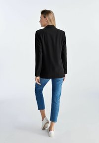 Czarna, dopasowana marynarka z pojedynczym rozcięciem z tyłu, noszona na jasnoniebieskie dżinsy kostiumowe i białe sneakersy, z długimi rękawami i ostrym kołnierzem.