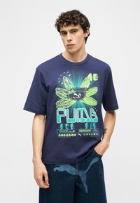 Νέος άνδρας με σγουρά μαλλιά φοράει μπλε σκούρο T-shirt Puma με μεγάλο σχεδιασμό από φλοράλ σε νέον πράσινο και λογότυπο της μάρκας, συνδυασμένο με αντίστοιχα σορτς.