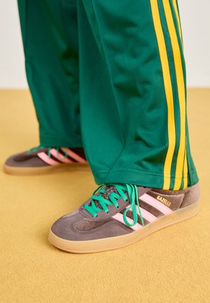 Osoba nosząca zielone spodnie dresowe z żółtymi paskami oraz szare buty Adidas Gazelle z różowymi paskami i zielonymi sznurówkami, stojąca na beżowej podłodze.