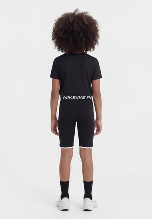 Dieťa s kučeravými vlasmi oblečené v čiernom šporotvom oblečení značky Nike, stojace chrbtom, na nohách má biele tenisky a čierne ponožky na bielom pozadí.