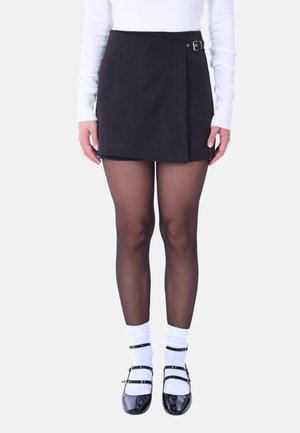 Gonna nera avvolgente con fibbia laterale, abbinata a collant neri trasparenti e scarpe nere in vernice. Il modello indossa una maglietta bianca a maniche lunghe.