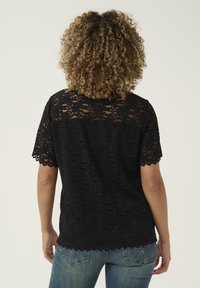 Black lace top with short sleeves, scalloped edges, and a floral pattern; paired with blue jeans, viewed from the back.
