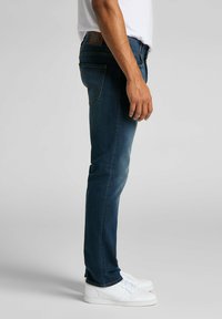 Tynde blå denimjeans med en let falming, prydet med et lædermærke i taljen og fem lommer. Matchet med hvide sneakers.