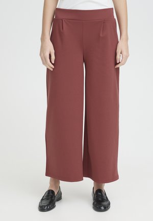 Donna che indossa pantaloni color ruggine a gamba larga e vita alta abbinati a mocassini neri, in piedi davanti a uno sfondo semplice.