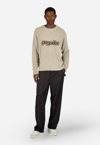 Krém színű bolyhos pulóver "Pagador" logóval; kerek nyakú; fekete, laza nadrággal és fehér tornacipővel viselve. A modell egy sima háttér előtt áll.