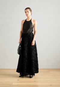 Haut halter noir associé à une jupe longue ornée d'accents floraux texturés. Tient un sac noir au design structuré.