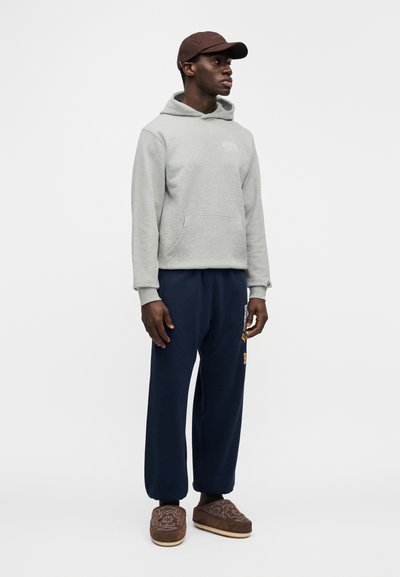 Γκρι φούτερ με κουκούλα και μπροστινή τσέπη, μπλε σκούρο παντελόνι φόρμας με κεντημένες λεπτομέρειες και καφέ παπούτσια τύπου slip-on με υφασματικά σχέδια, ολοκληρωμένο με καφέ καπέλο.