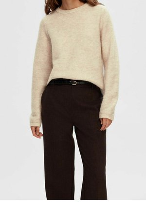 Vrouw die een crèmekleurige gebreide trui draagt, ingestopt in een hoge taille donkerbruine broek met een zwarte riem.