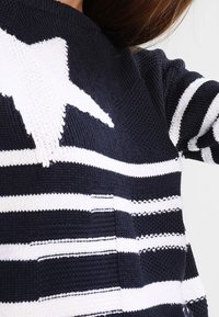 Pull tricoté bleu marine avec des rayures horizontales blanches et un grand motif étoile. Tissu texturé avec un col rond et des manches longues.