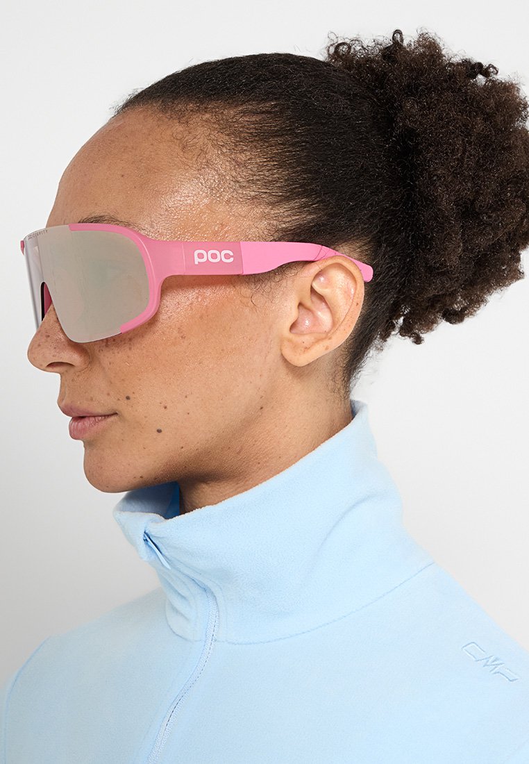 Kobieta z kręconymi włosami w kucyku, nosząca różowe okulary przeciwsłoneczne w stylu wraparound oraz jasnoniebieską polarową kurtkę z wysokim kołnierzem.