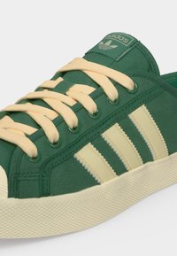 Πράσινο παπούτσι Adidas με μπεζ κορδόνια, μπεζ σόλα και τρεις μπεζ ρίγες στο πλάι με το λογότυπο Adidas στη γλώσσα.