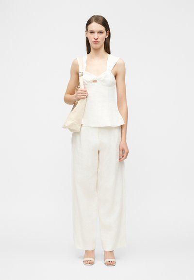 Γυναίκα που φοράει λευκό αμάνικο τοπ και παντελόνι με φαρδιά πόδια, κρατώντας μπεζ τσάντα ώμου, στέκεται μπροστά σε απλό φόντο.