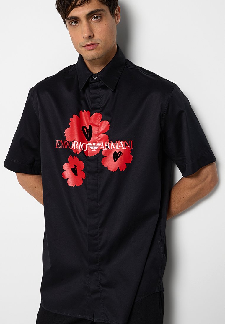 Joven con una camisa negra de manga corta con diseños florales y de corazones rojos y el logo de "Emporio Armani" en el frente.