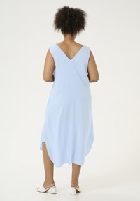Helles blaues ärmelloses Kleid mit V-Ausschnitt, seitlichen Schlitzen und lockerer Passform, hergestellt aus weichem Stoff. Kombiniert mit weißen Absatzsandalen.