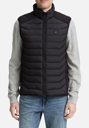 Schwarze Steppweste mit hohem Kragen und Reißverschluss vorne, gesteppte Struktur und Logopatch; getragen über einem grauen Langarmshirt und Jeans.