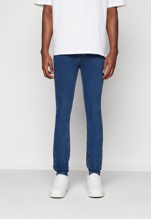 Blue denim skinny jeans with a smooth texture, featuring five pockets and a zip-fly closure, paired with white sneakers and a loose-fitting white t-shirt.