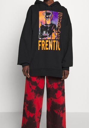 Osoba nosząca oversize czarną bluzę z kapturem z pomarańczową i fioletową grafiką i tekstem, zestawioną z szerokimi czerwono-czarnymi spodniami tie-dye.