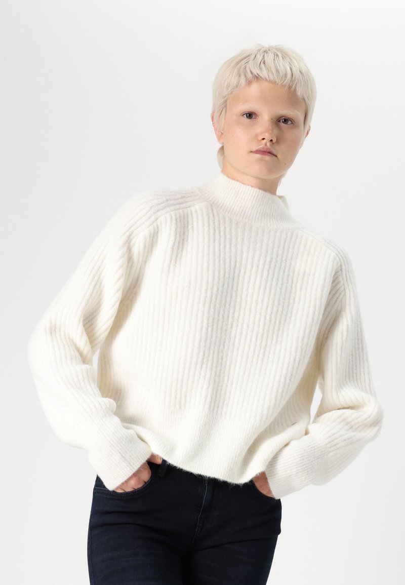 Maglione bianco a coste con colletto alto, vestibilità oversize e maniche larghe, realizzato in materiale morbido e strutturato. Indossato con jeans scuri.