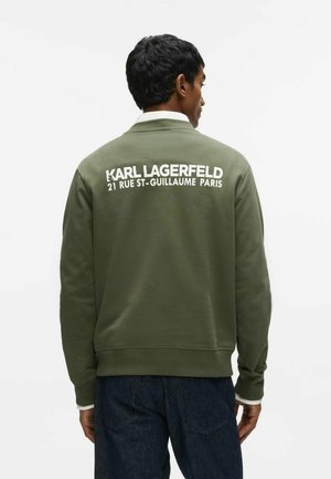 Homme portant un sweat-shirt vert olive avec un texte blanc "KARL LAGERFELD 21 RUE ST-GUILLAUME PARIS" dans le dos, jean foncé.