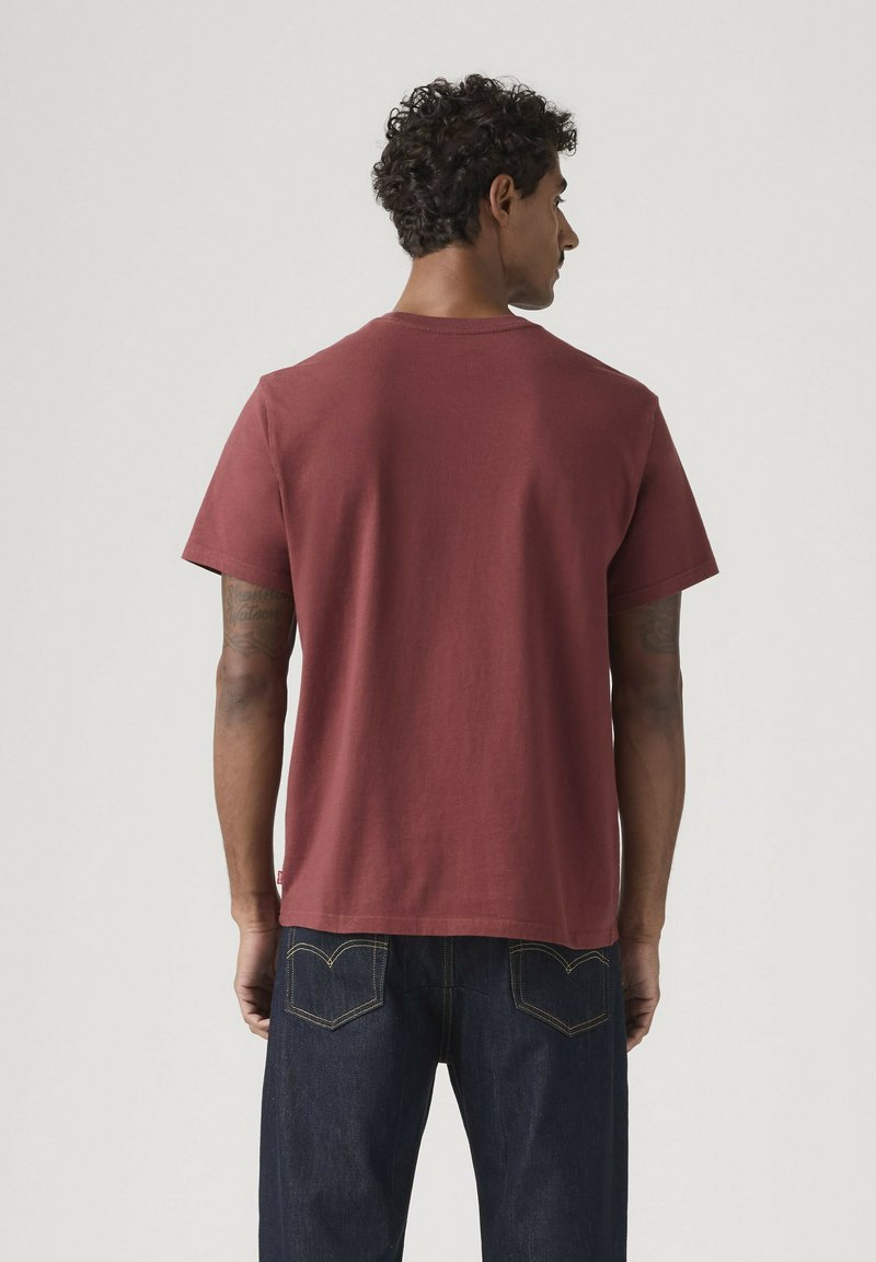 Mand med krøllet hår set bagfra iført en ensfarvet mørkerød kortærmet t-shirt og mørkeblå jeans mod en ensfarvet baggrund.