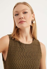 Top senza maniche di colore verde oliva con un motivo a zigzag testurizzato. Orecchini dichiarativi in oro con un design ondulato. Sfondo semplice e neutro.