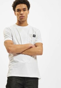 Hvid bomulds T-shirt med rund halsudskæring, korte ærmer og en venstre brystlomme med et sort mærke-logo. Model i en neutral positur.