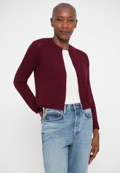 Cardigan de malha bordeaux com um padrão texturizado, gola redonda, bolsos frontais e fecho de botão, usado sobre uma blusa branca e jeans azuis.