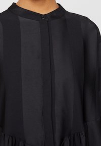 Närbild av en svart skjorta med vertikala ränder, en rund krage och en enda svart knapp vid halsen.