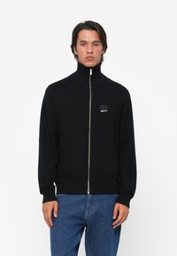 Jauns vīrietis, kurš valkā melnu zip-up džemperi ar augstu apkakli un zilo džinsu bikses, stāvot uz balta fona.