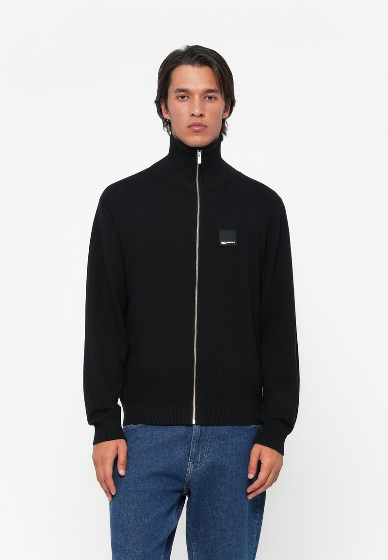 Jauns vīrietis, kurš valkā melnu zip-up džemperi ar augstu apkakli un zilo džinsu bikses, stāvot uz balta fona.
