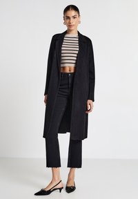 Cappotto lungo nero realizzato in un tessuto morbido, indossato sopra un top corto a righe. Jeans neri a vita alta con orli sfrangiati e tacchi slingback neri.
