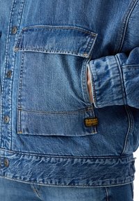 Jeansjacka i blå ton, med två bröstfickor, knappstängning och G-Star-logotyp. Tyget visar en lätt textur och sömnadsdetaljer.