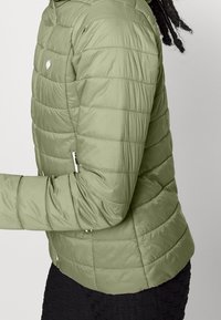 Personne portant une veste matelassée olive avec des poches zippées, montrée de profil contre un fond clair.