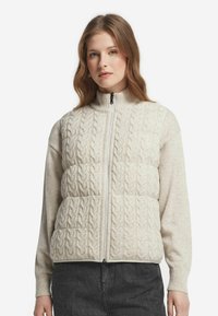 Junge Frau mit hellbraunem Haar, die eine cremefarbene, kabelstrickartige, zip-up Weste über einem beigen Langarmpullover und dunklen Jeans trägt.