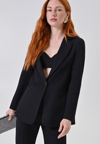 Blazer nero su misura con rever a punta e due tasche frontali, abbinato a un top corto nero. Accessoriato con una pochette argentata.