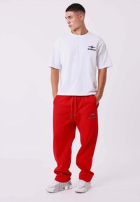 Joven con pelo corto, llevando una camiseta gráfica blanca, pantalones deportivos rojos holgados y zapatillas blancas, de pie con las manos en los bolsillos, mirando hacia la izquierda.