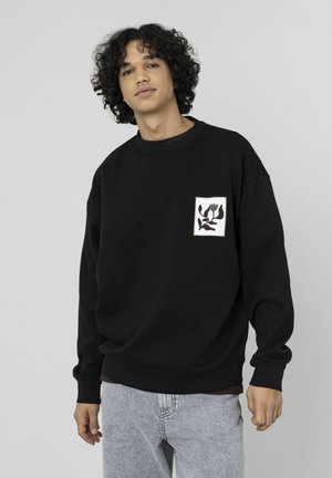 Sweatshirt noir oversize en coton, avec un patch floral carré sur la poitrine gauche et des poignets et un ourlet côtelés. Un jean gris complète le look.