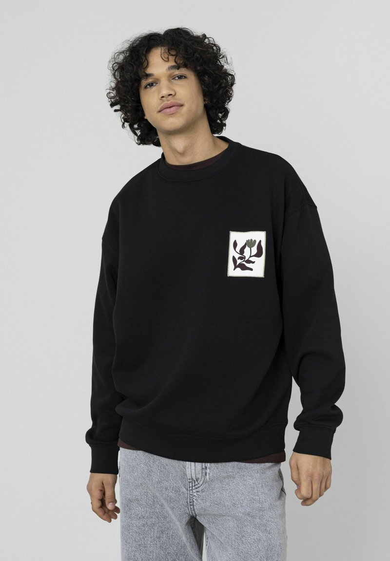 Sweatshirt noir oversize en coton, avec un patch floral carré sur la poitrine gauche et des poignets et un ourlet côtelés. Un jean gris complète le look.