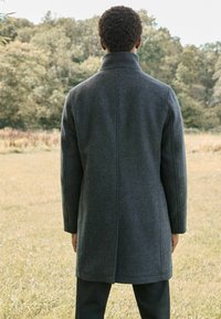 Abrigo de lana gris con cuello alto y corte recto, que presenta bolsillos laterales discretos y costuras visibles a lo largo de los dobladillos.