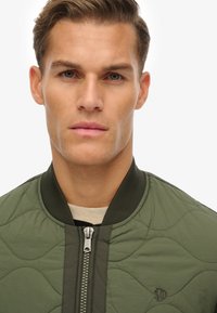 Groene gewatteerde bomberjack met een rits aan de voorkant en een geribde kraag, met een subtiele logo op de borst en een gladde textuur.