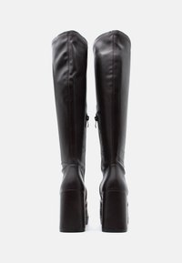 Bottes montantes en cuir marron avec une finition brillante, talons carrés, et détails de couture visibles aux coutures, dotées d'une fermeture éclair.