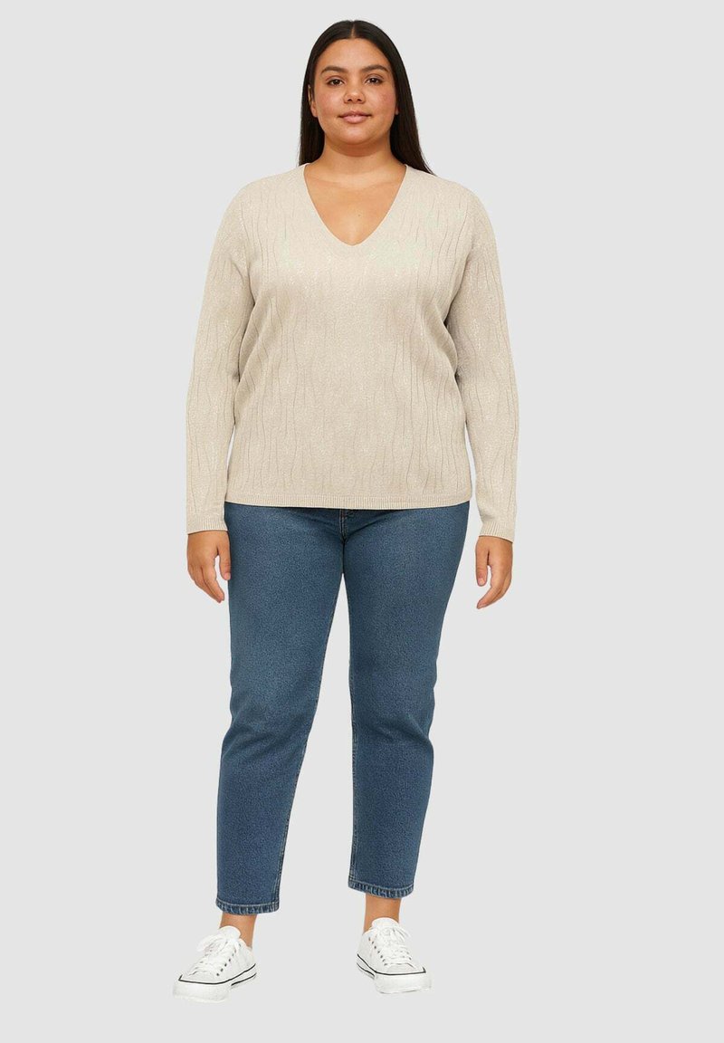 Maglione beige a maglia con scollo a V, caratterizzato da un motivo a onda testurizzato. Abbinato a jeans blu e sneakers bianche.