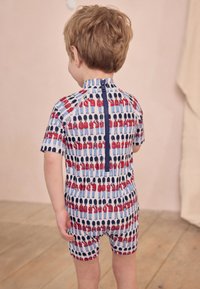 Kinder-Badeanzug mit kurzen Ärmeln, blauem Grundton und Muster mit Spielzeugsoldaten in Rot und Blau. Verfügt über einen Reißverschluss auf der Rückseite.
