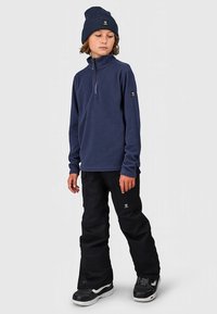 Πουλόβερ από ναυτικό φλίς με φερμουάρ στο μέσο, συνδυασμένο με μαύρο παντελόνι χιονιού. Φορεμένο με ταιριαστό ναυτικό σκουφί και μαύρα παπούτσια με λευκές σόλες.