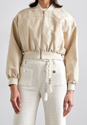 Femme portant une veste beige zippée avec des poignets élastiques et un pantalon blanc texturé avec des poches devant et une ceinture tressée à pampilles.