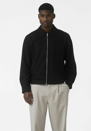 Sort lynlåsjakke med krave, der har subtile nålestriber og en lige pasform. Kombineret med lysegrå, plisserede bukser.