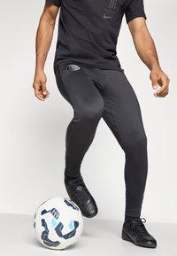 Sorte atletiske leggings med tettsittende passform, med en logo på venstre lår, kombinert med svarte fotballsko og en mønstret hvit fotball.