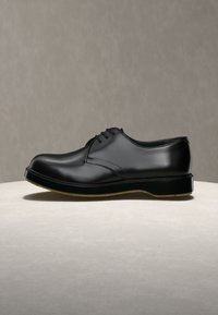 Γυαλιστερό μαύρο δερμάτινο ανδρικό επίσημο παπούτσι με κορδόνια και παχύ πάτο, εκτεθειμένο σε ουδέτερη ανοιχτή επιφάνεια με φόντο σε απαλό γκρι χρώμα.