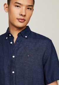 Camicia a maniche corte di colore blu navy realizzata in tessuto testurizzato con colletto a choker, con un piccolo logo sul petto e bottoni bianchi.